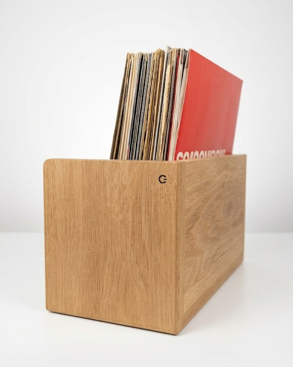 A light oak wooden record box with a visible grain, holding several vinyl records including a prominent red sleeve, set against a clean white studio background.
