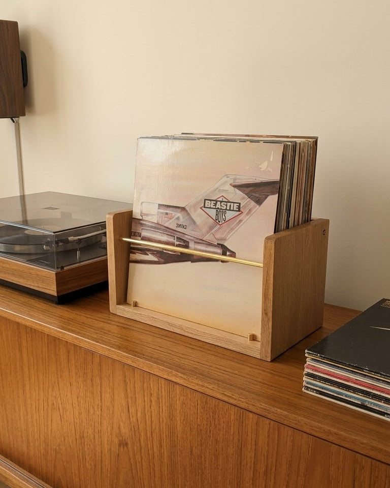 A light natural wood vinyl record holder with a slatted front, holding several records, placed on a mid-century modern wooden sideboard next to a turntable.
