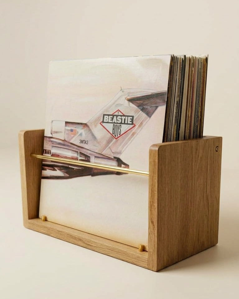 A light oak wood vinyl record holder with a gold metal bar, holding a record album and several records, set against a warm beige background.