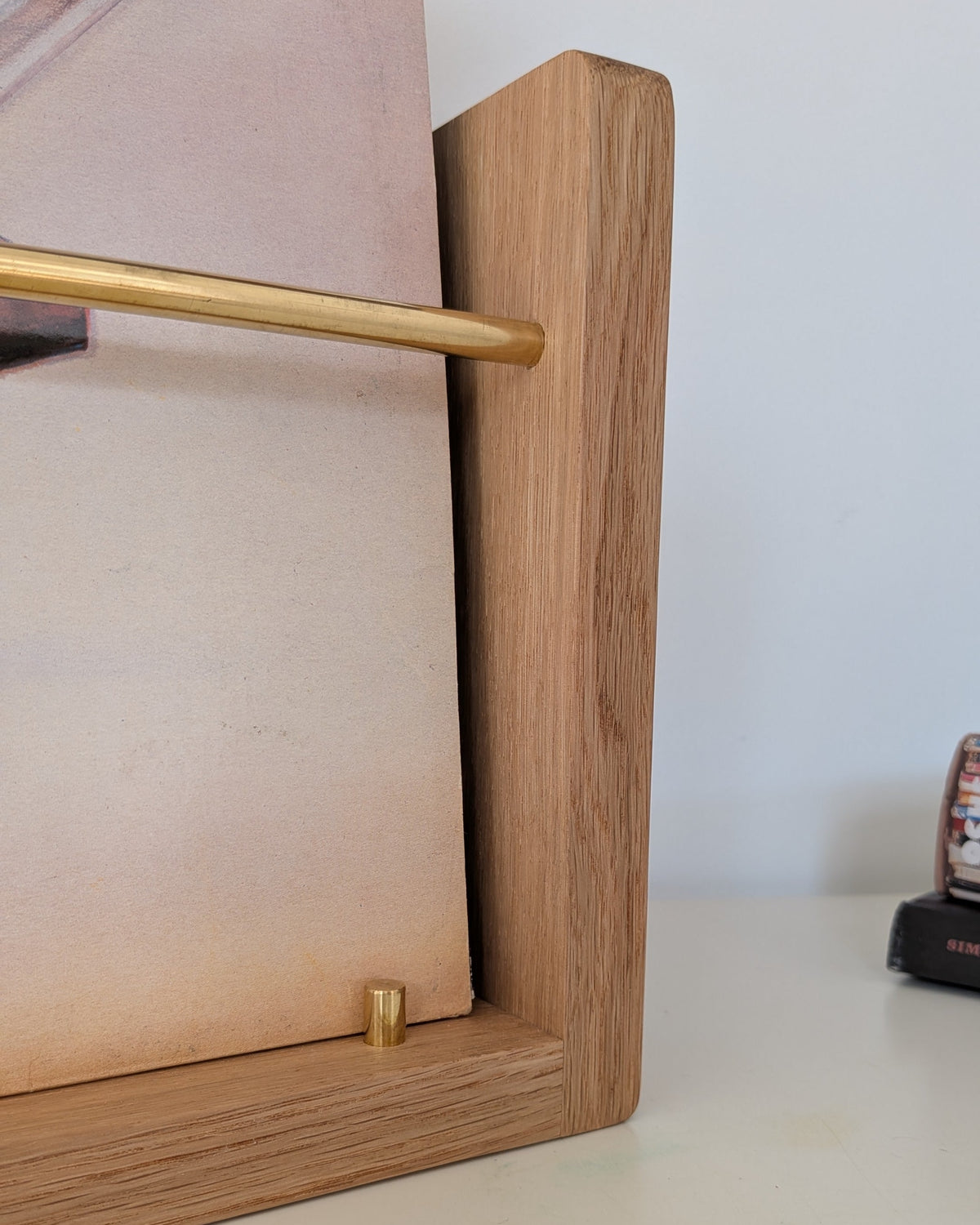 Close-up of a wooden picture frame with a gold metal rod and gold mounting hardware, shown on a light-colored tabletop against a white wall.