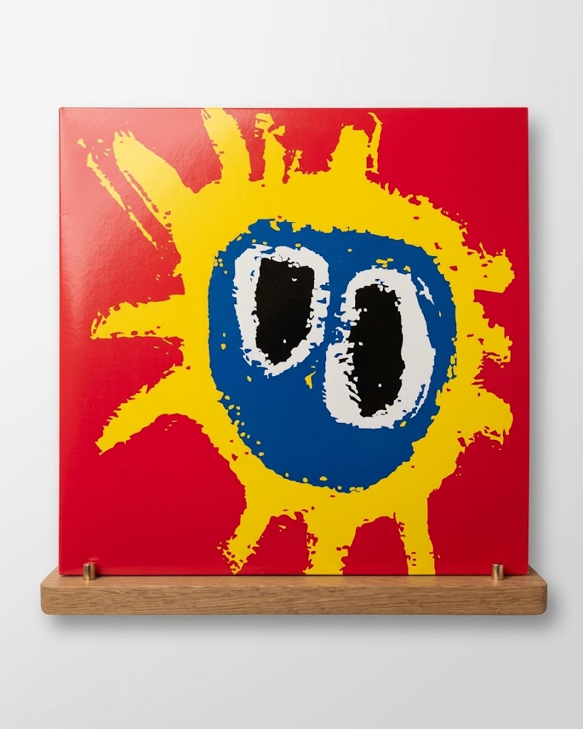 A square abstract art print or canvas mounted on a wooden display stand against a light gray wall. The artwork features a bright red background with a large yellow paint-splatter sun shape and a central blue circle containing two black-and-white oval eyes.