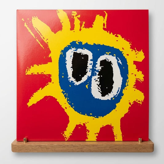 A square abstract art print or canvas mounted on a wooden display stand against a light gray wall. The artwork features a bright red background with a large yellow paint-splatter sun shape and a central blue circle containing two black-and-white oval eyes.