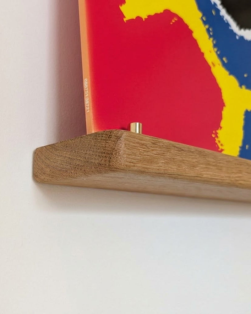 Close-up of a wooden floating shelf mounted on a white wall, holding the lower edge of a colorful abstract art print with red, yellow, and blue brushstrokes.