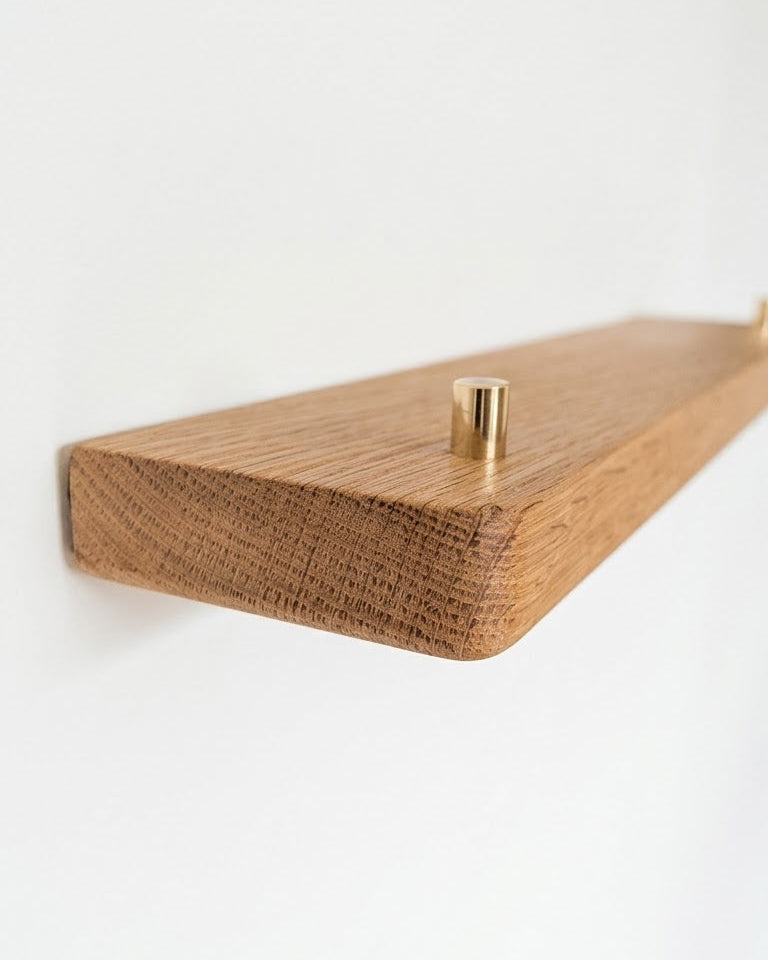 A minimalist floating wooden shelf with a warm medium-brown wood grain finish and a small gold metal peg, shown mounted on a plain white wall.