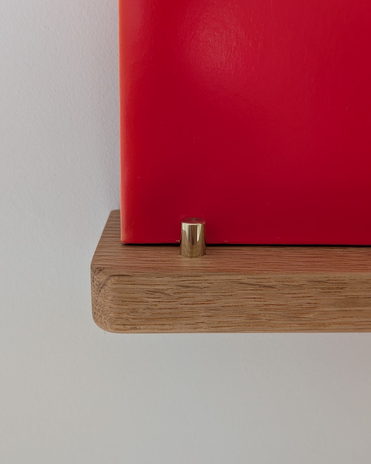 A close-up of a red wall-mounted shelf or panel with a small brass cylindrical hardware piece at the bottom, mounted on a light wood shelf board against a plain light gray wall.