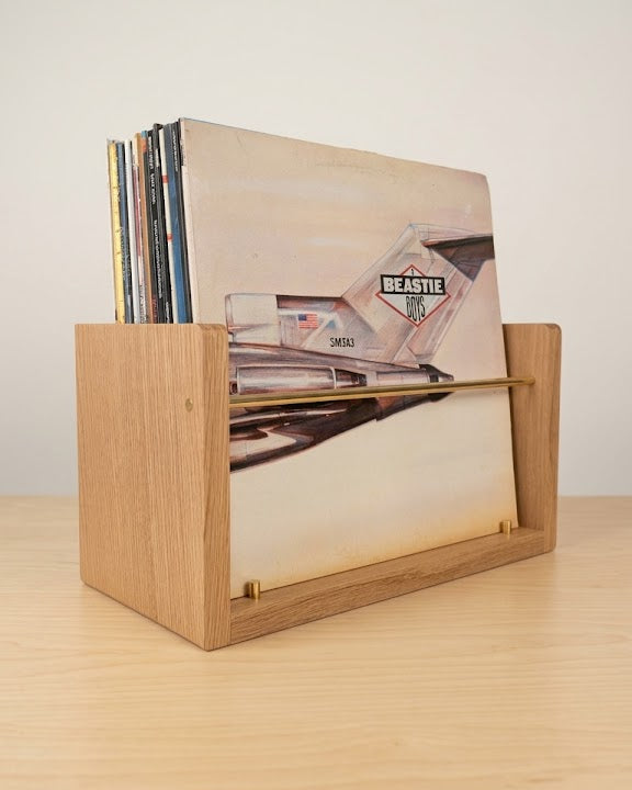 A light oak wooden record holder with a slatted front, holding several vinyl records, set on a light wood tabletop against a neutral off-white background.