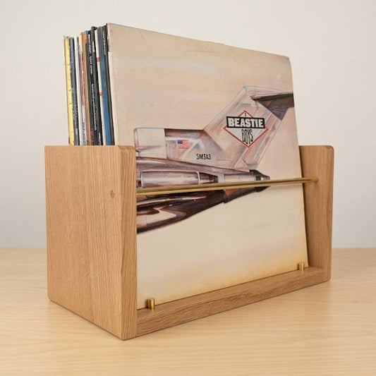 A light oak wooden record holder with a slatted front, holding several vinyl records, set on a light wood tabletop against a neutral off-white background.