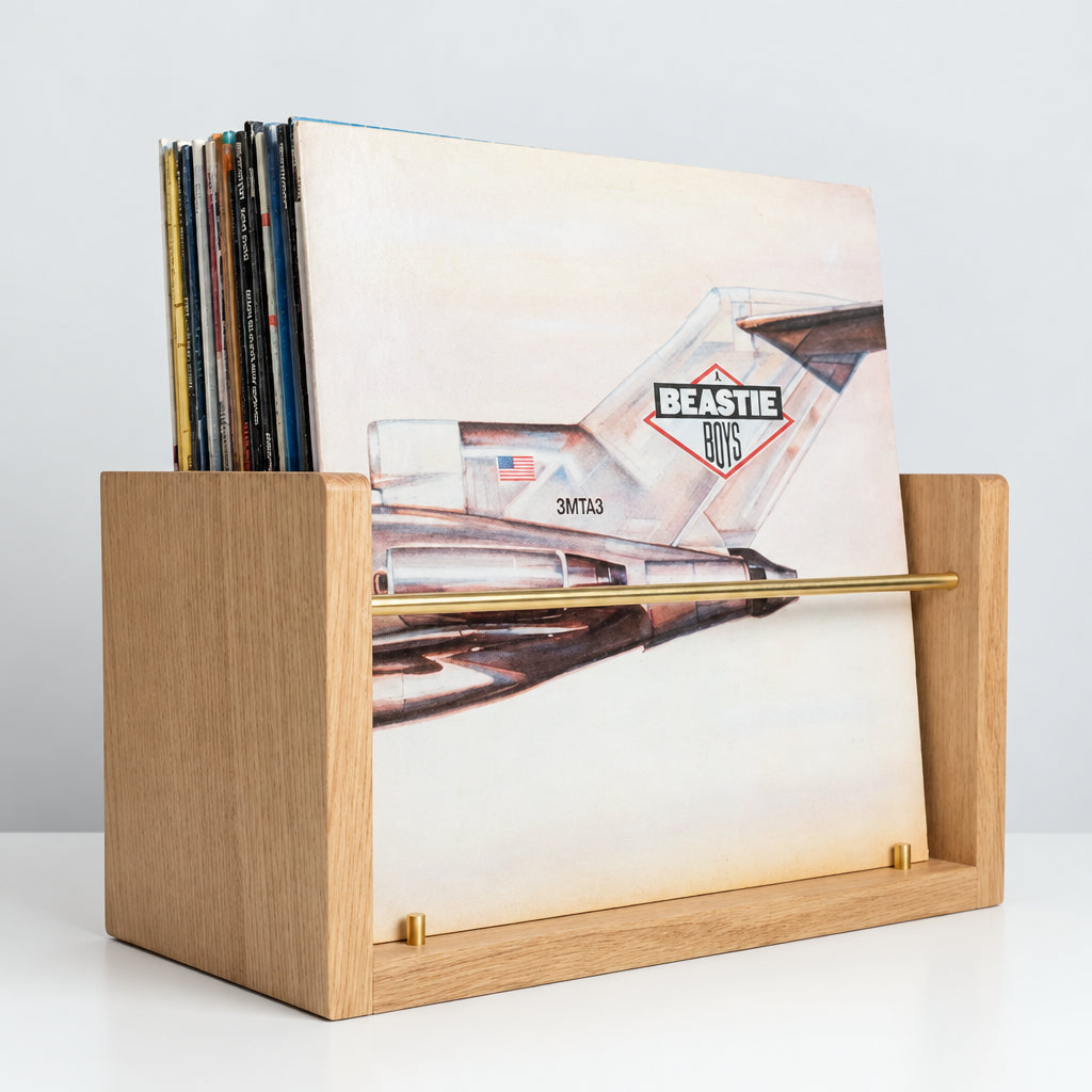 A light oak wood vinyl record holder with a gold metal guide bar, holding several records including a visible Beastie Boys album, set against a neutral gray background.