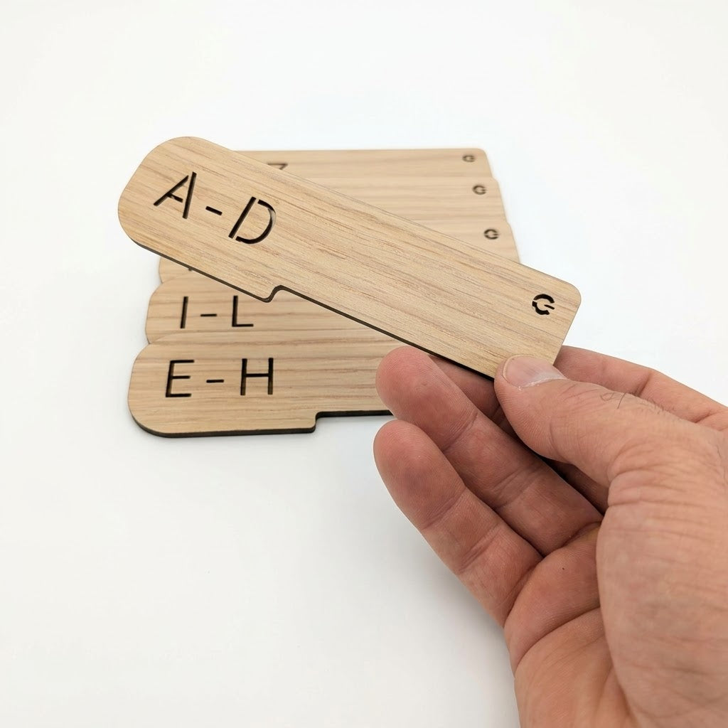 A hand holding a light natural wood magnetic letter sorter card showing 'A-D', with other cards displaying 'I-L' and 'E-H' stacked behind it on a white background.